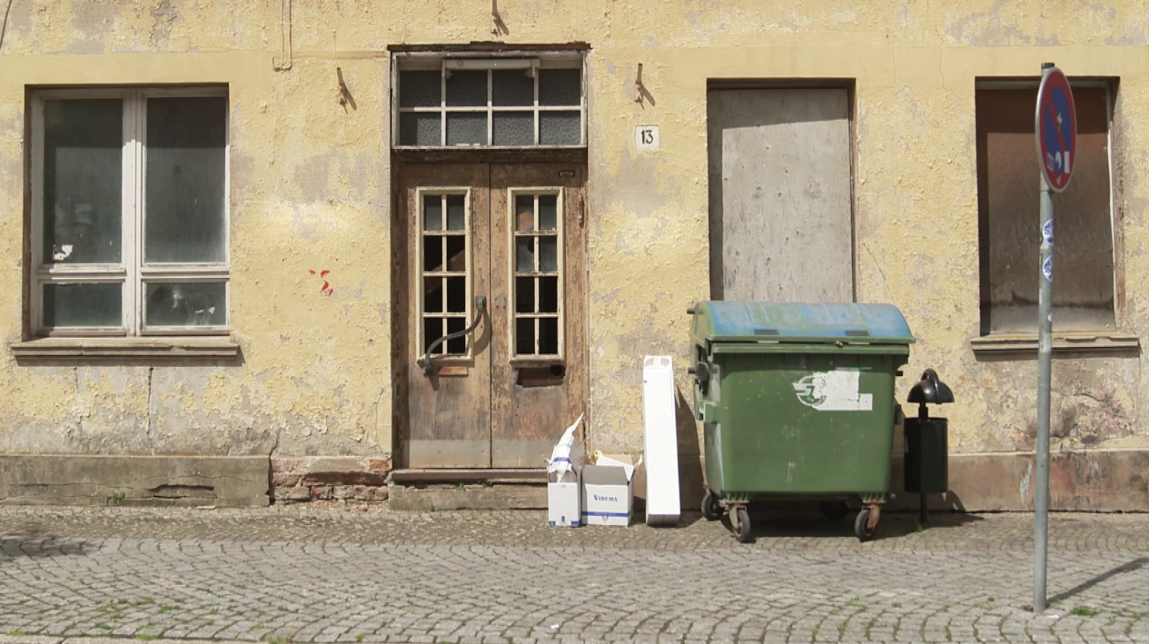 Eine alte Hauswand mit schmutzigen und abgedeckten Fenstern. Davor stehen zwei Mülltonnen, einige Kartons und ein Straßenschild.